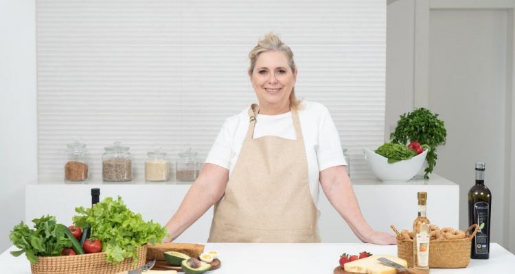 A mature woman in an apron stands at a kitchen counter with fresh produce and cheese, ready for cooking.